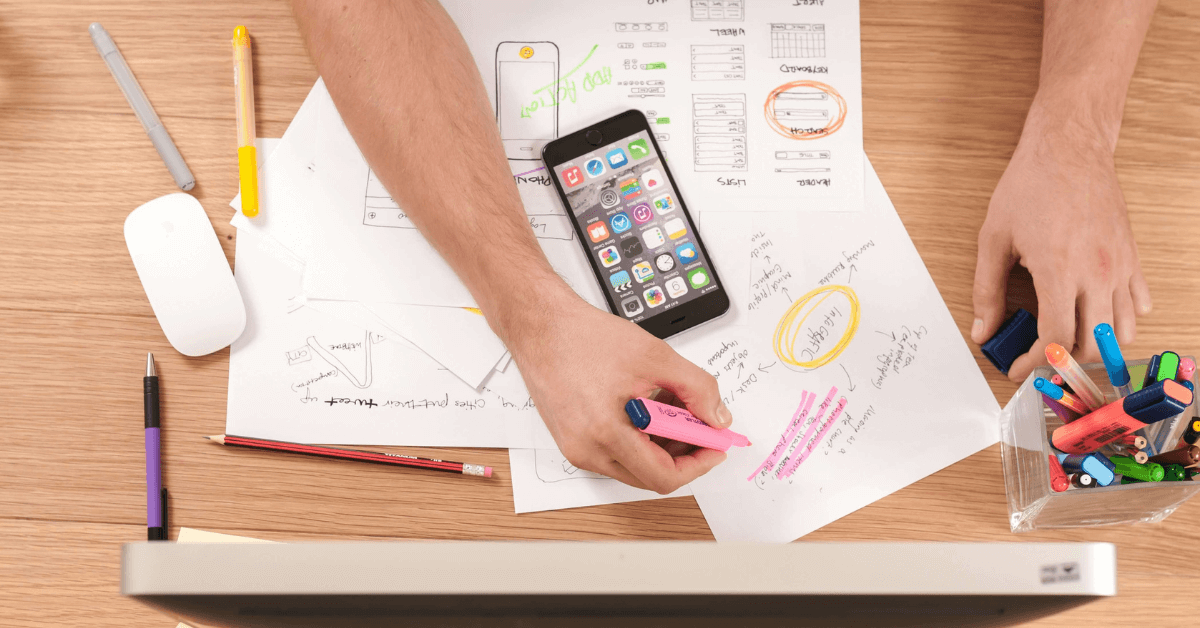 An overhead view of a workspace with a smartphone, papers with sketches and annotations, and a variety of colorful markers, potentially representing a resource for Japanese language learners.