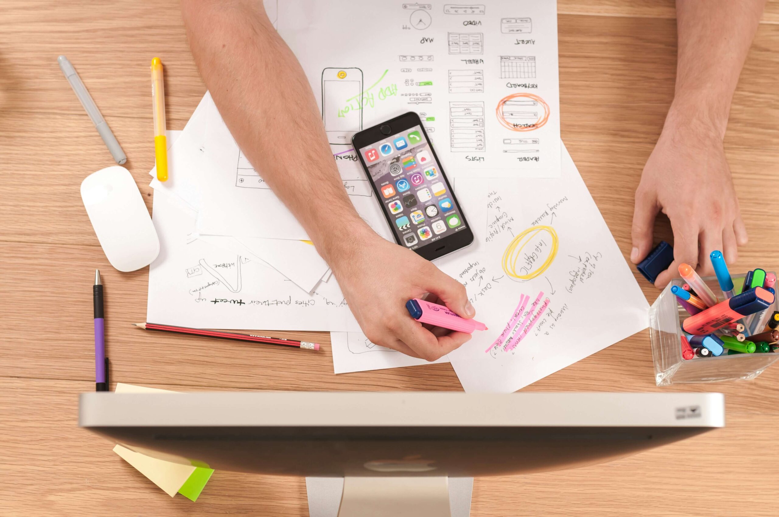 An overhead view of a workspace with a smartphone, papers with sketches and annotations, and a variety of colorful markers, potentially representing a resource for Japanese language learners.
