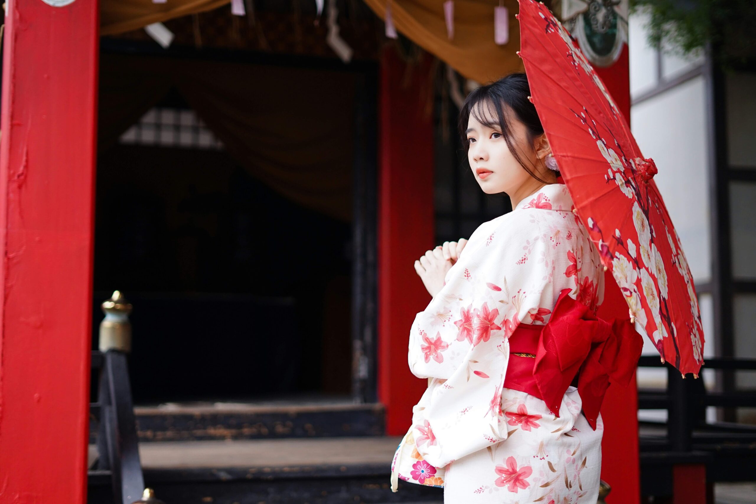 A woman in a kimono with cherry blossom designs holding a red umbrella, standing at the entrance of a traditional Japanese building.