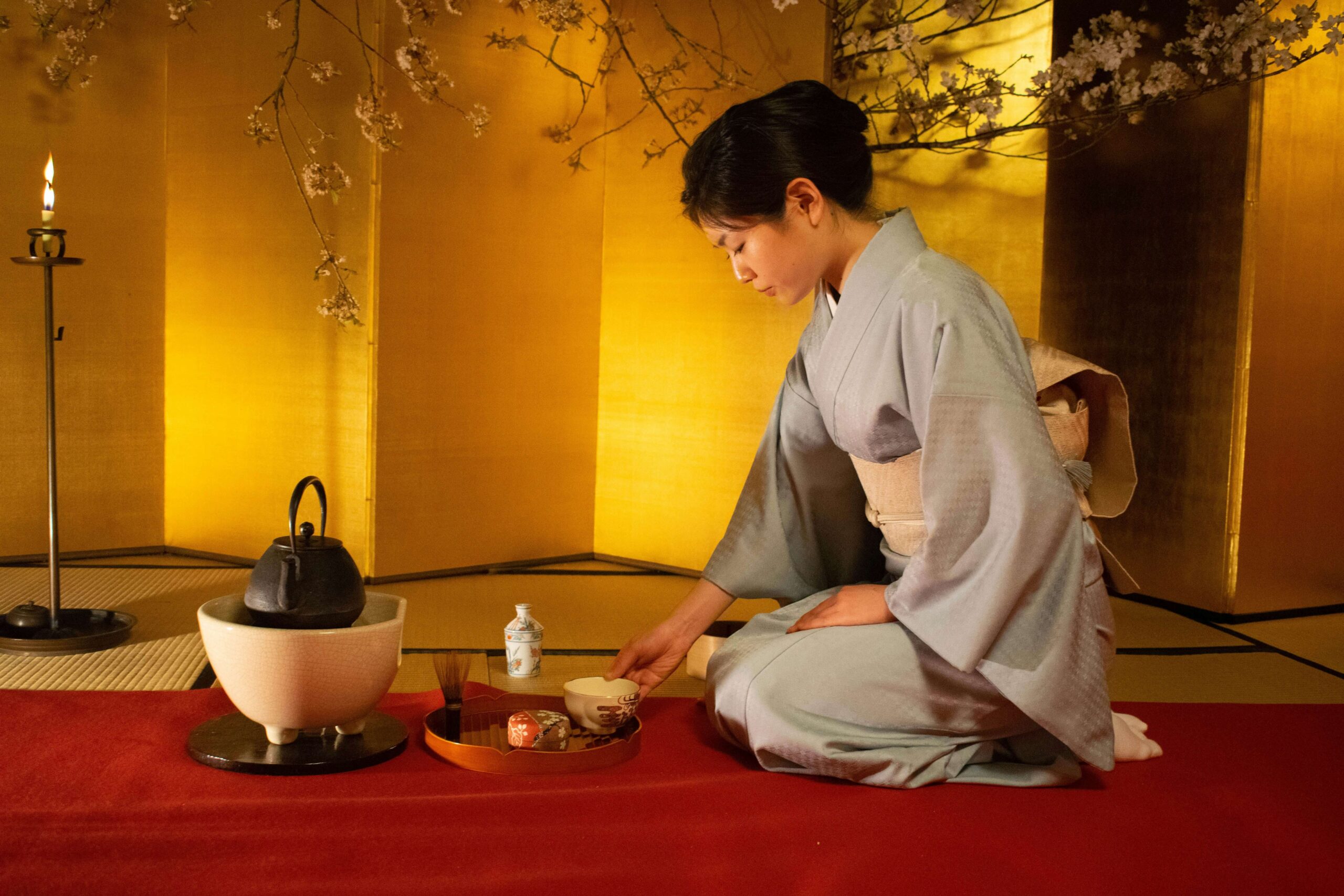 A woman in a traditional Japanese kimono is performing a tea ceremony, with a focus on preparing tea utensils on a red mat in a room with a golden sliding door and cherry blossoms.
