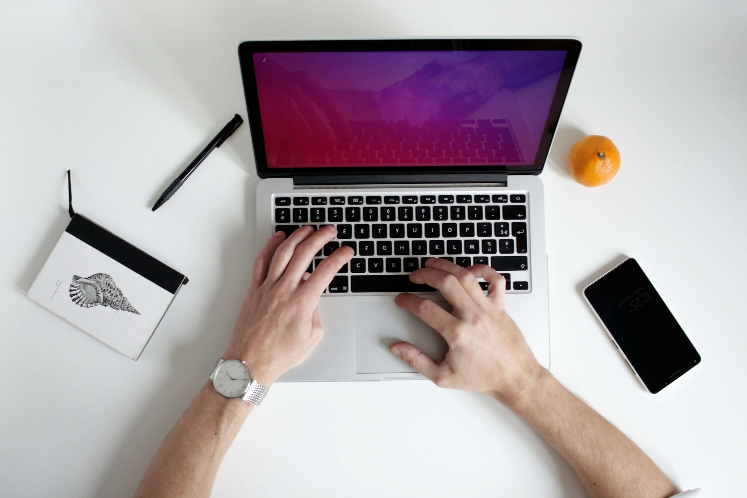 Overhead view of a person typing on a laptop, possibly using Romaji input, with a notebook, pen, smartphone, and an orange placed on a white desk.