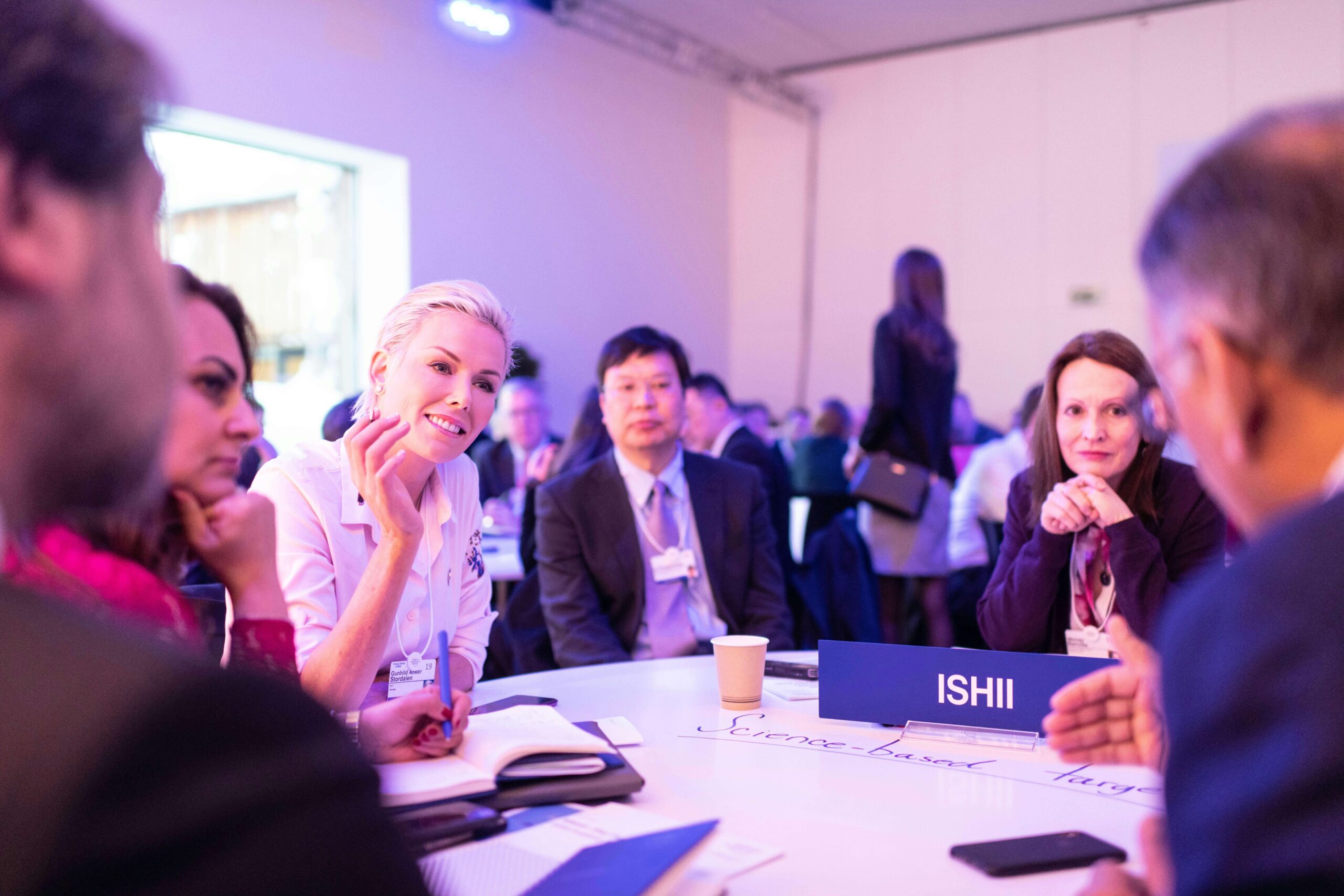 This image depicts a diverse group of professionals engaging in a discussion at a conference table, which can illustrate the use of honorific titles in a formal, multicultural business setting. One of the name tags visible says "ISHII," suggesting that honorific titles may be addressed or referenced in the conversation.