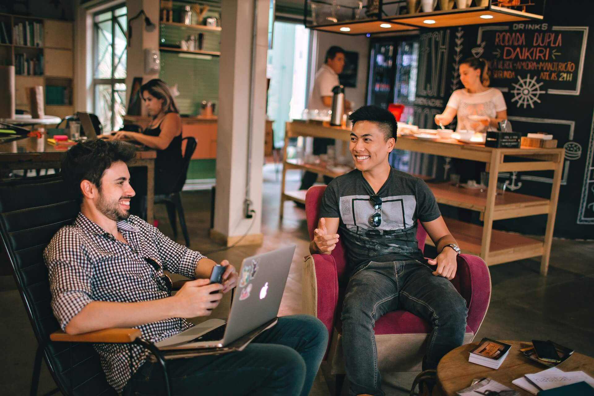 This image shows two young men talking and smiling in a cozy café. One is using a laptop, and others are in the background. The image is for a blog about the use of "-ne" and "-yo" in sentences.
