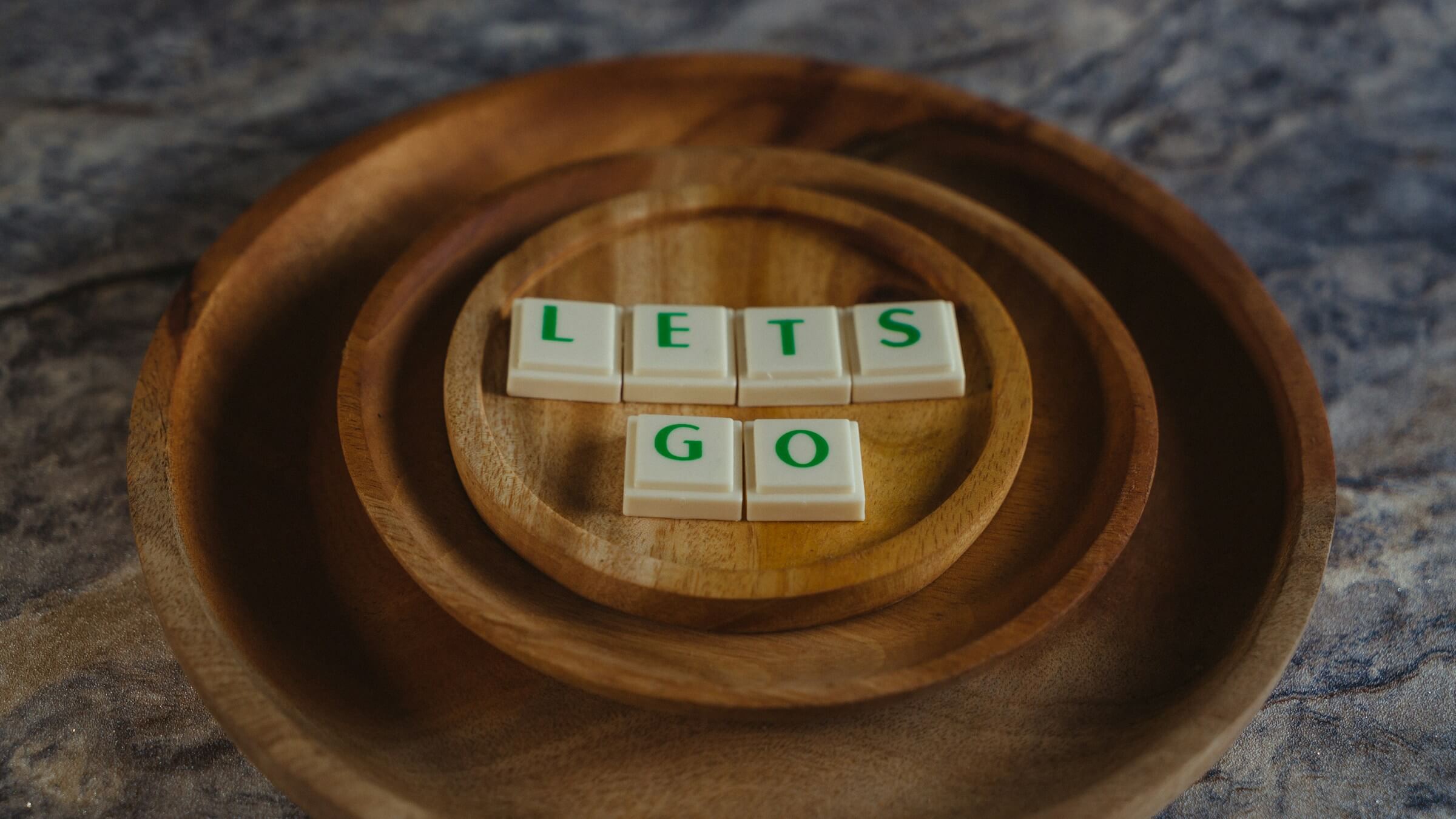 A set of wooden bowls arranged in concentric circles, with letter tiles spelling out "LETS GO" placed in the center. This image represents the Japanese grammatical concept -ましょう (mashou), used to suggest or propose doing something together, similar to saying "let's" in English.
