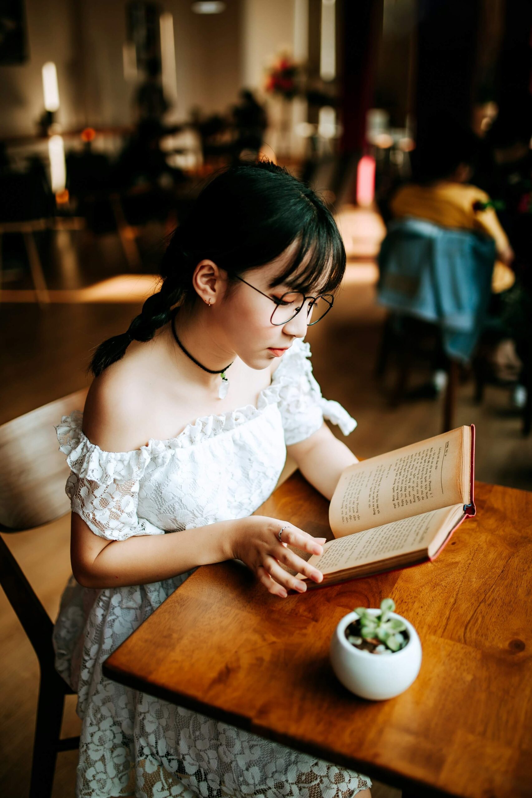 A young woman wearing glasses and a white dress is sitting at a table reading a book. This image represents the concept of -ている (-ている, teiru) to describe an action in progress in Japanese.
