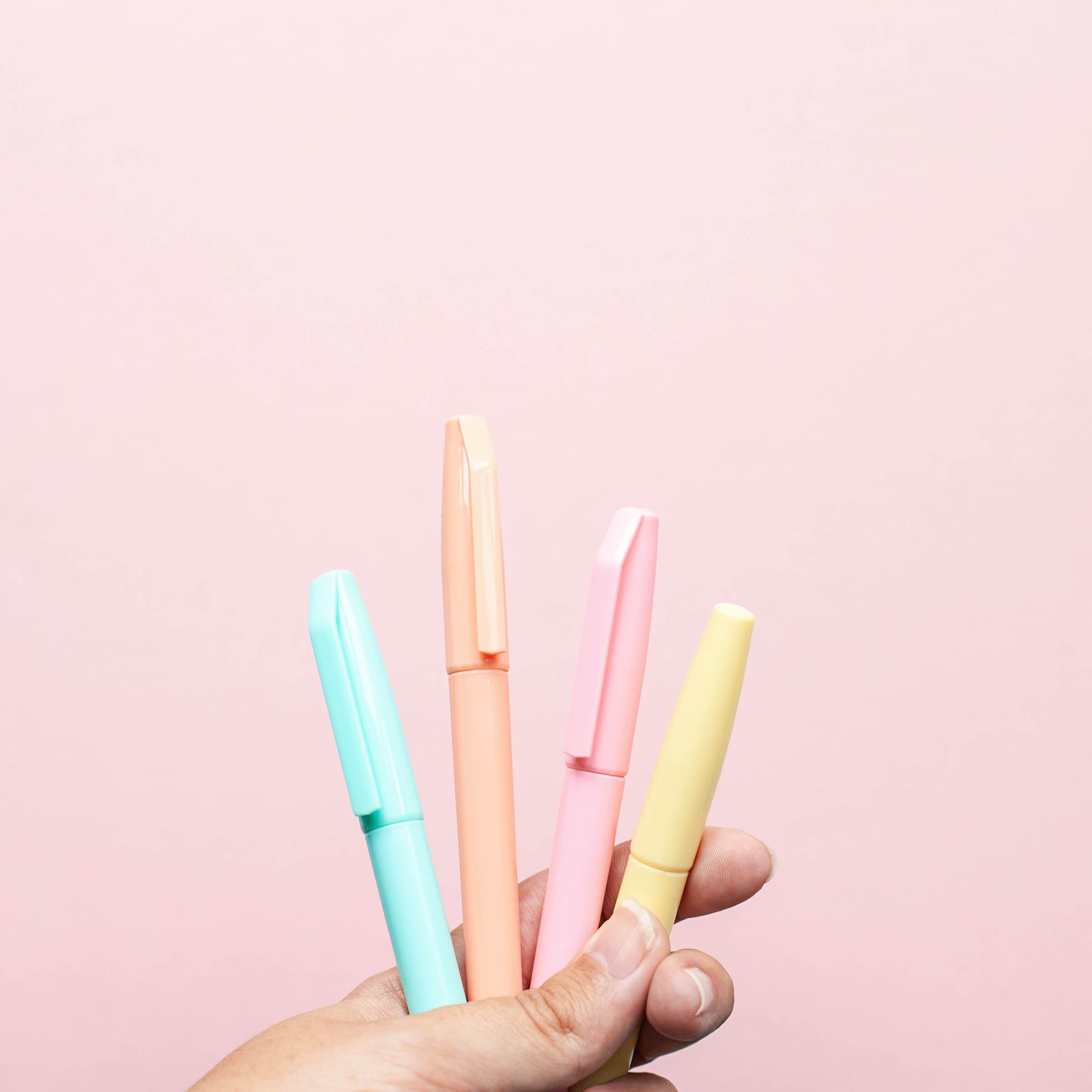 A hand holding four colorful pens against a pink background. This image represents the Japanese counter for long objects, such as pens, using the counter 本 (ほん, hon).
