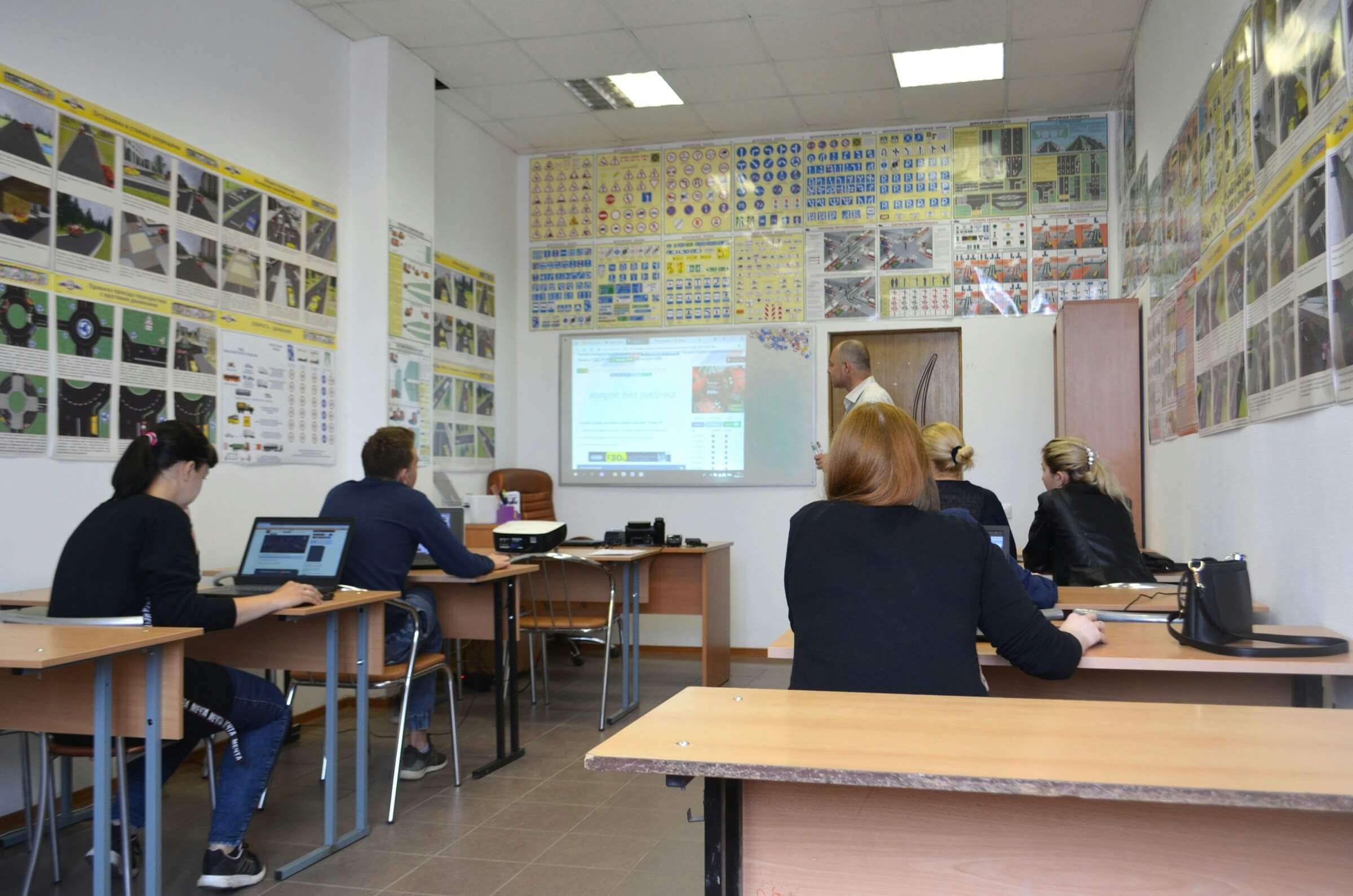 Students in a classroom listening to a teacher's explanation. The walls are covered with numerous posters. This image represents "Counting People (There are - people in the classroom).”
