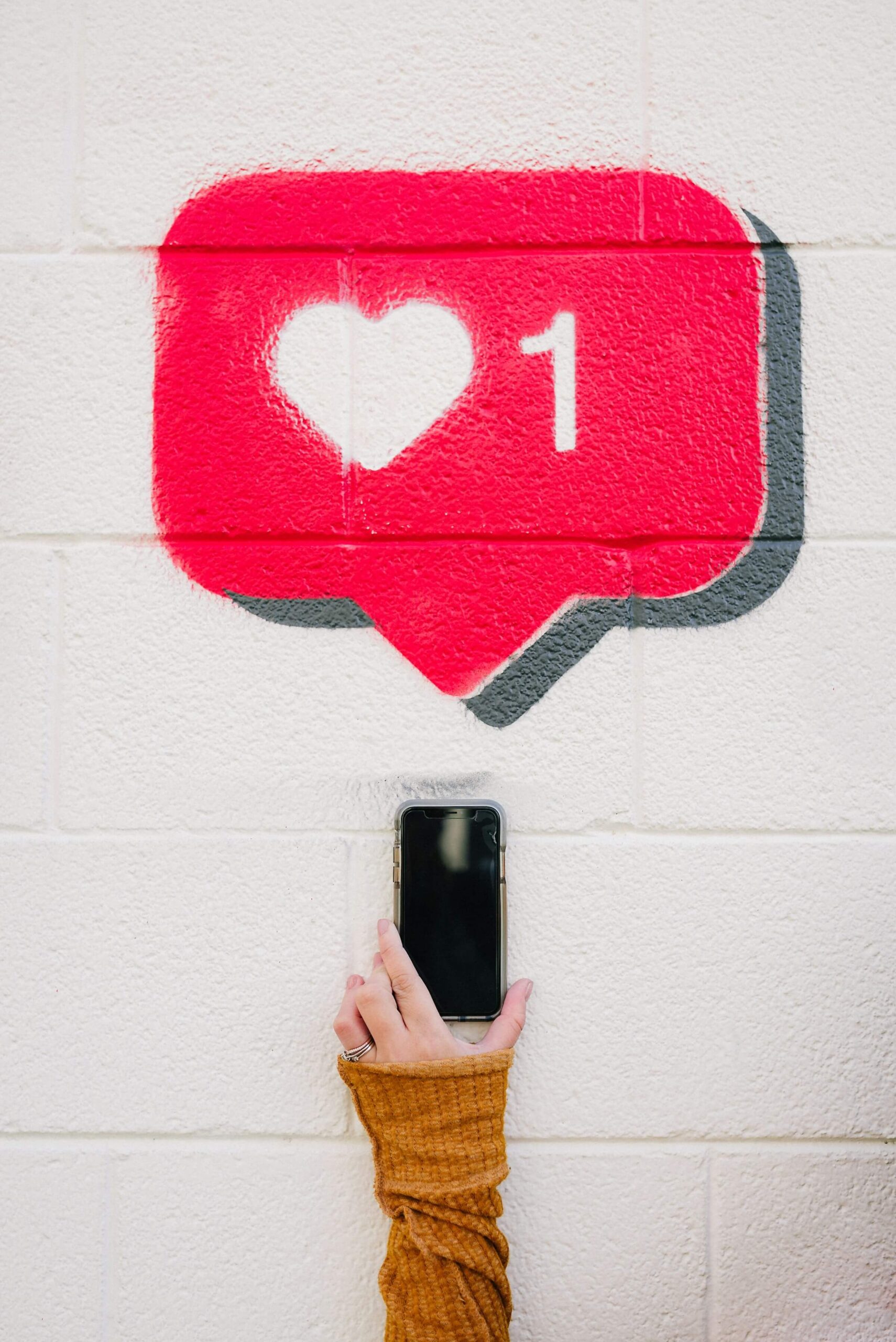 An image showing a person's hand holding a smartphone against a white wall. Above the phone is a large, red heart icon with the number 1, resembling a social media "like" notification. This image represents the concept of "liking" or "being fond of" something, which aligns with the Japanese adjective "好きな" (sukina).
