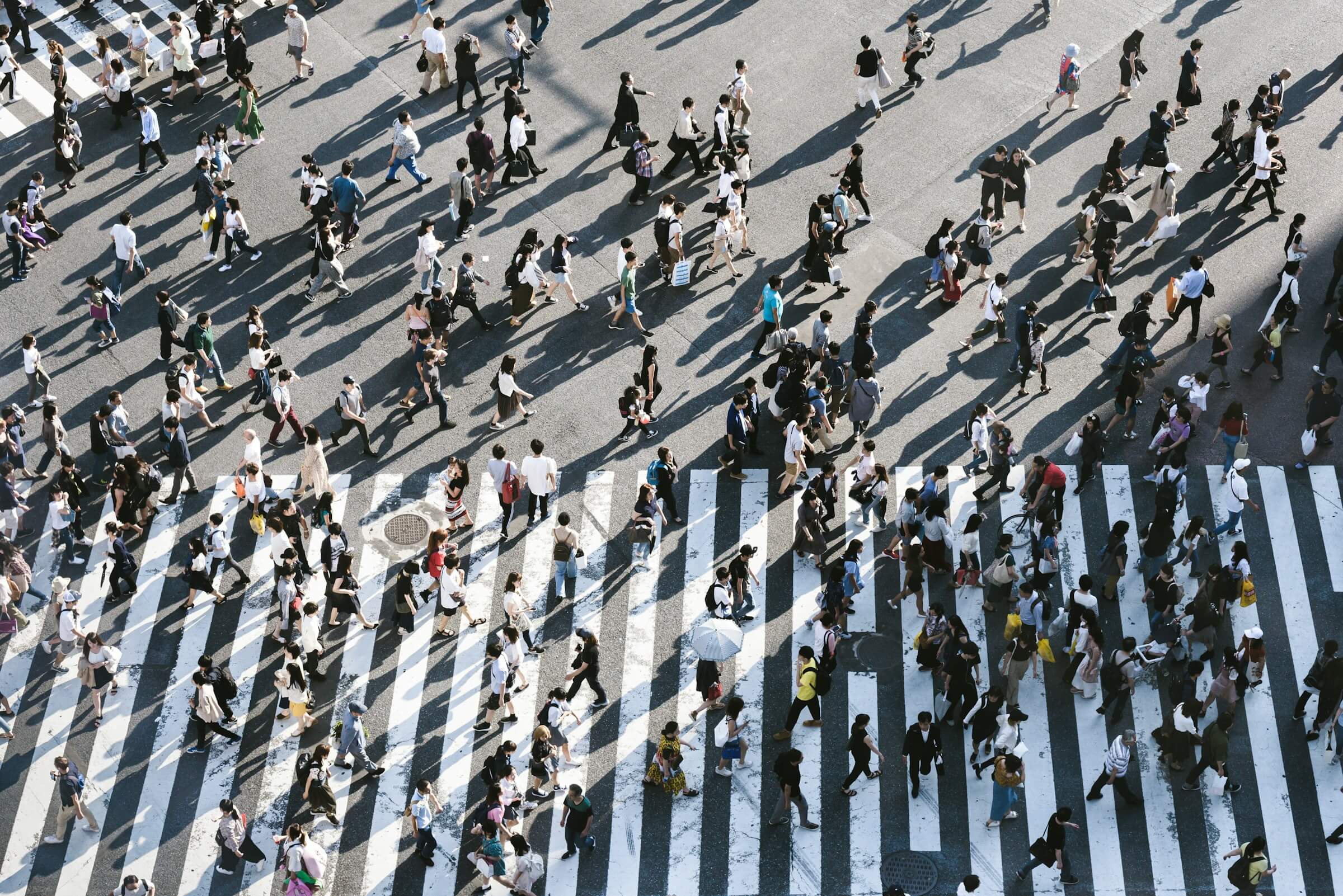 An aerial view of a busy crosswalk filled with many people walking in various directions, illustrating the phrase "人がたくさんいます" (Hito ga takusan imasu). 