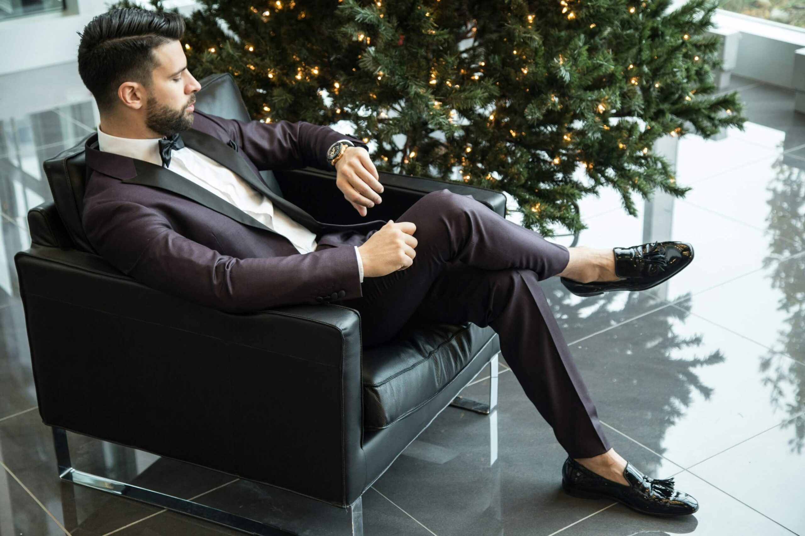 A man dressed in a suit is sitting relaxed in a chair, looking at his watch. He is in a modern, well-lit space with a decorated Christmas tree in the background. This image represents "-時間" (hours).
