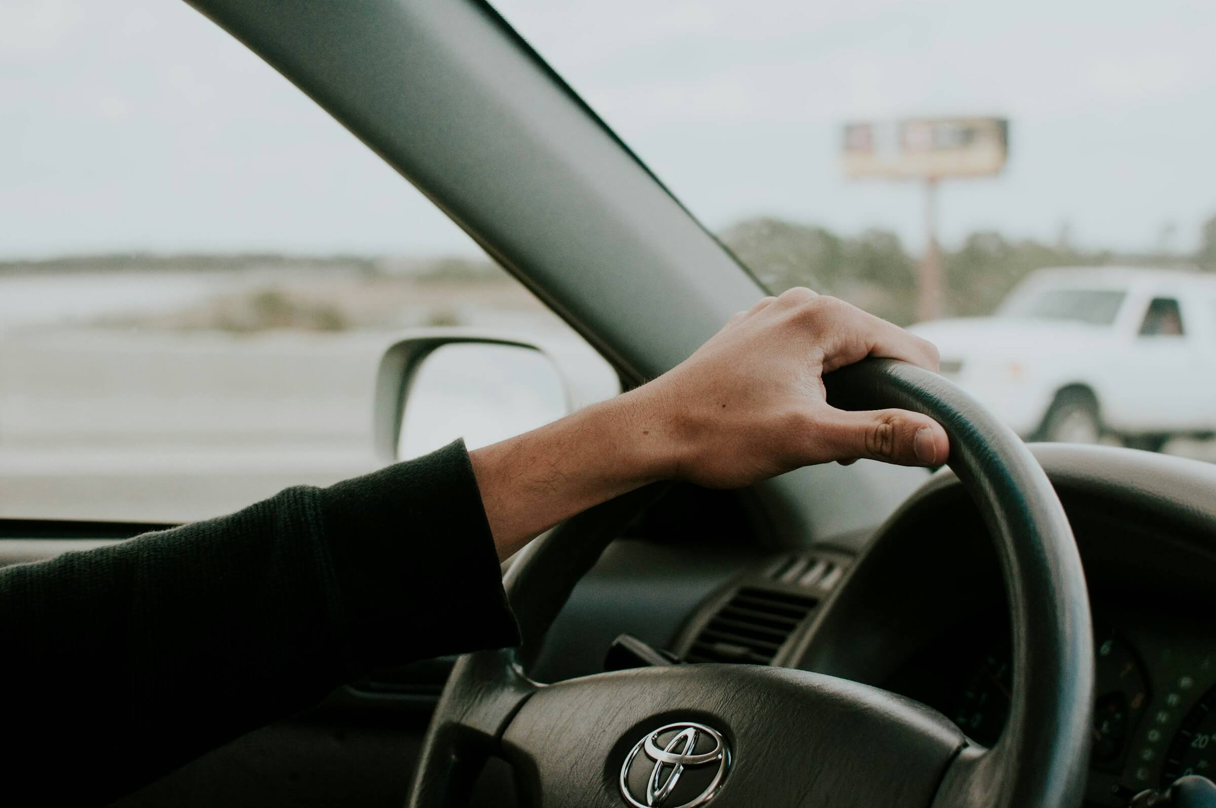 This image shows a person's hand on a car steering wheel, illustrating the concepts of 'ドライブ' (doraibu) for leisure driving and '運転する' (unten suru) for general driving in Japanese.
