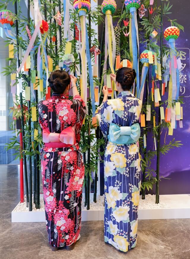 This image shows two women in colorful kimonos hanging tanzaku (wish papers) on bamboo branches during the Tanabata festival. The scene captures the vibrant and traditional celebration of this Japanese festival, where participants write wishes and hang them on bamboo, hoping for their dreams to come true. The backdrop features a thematic decoration of bamboo and stars, enhancing the festive atmosphere of Tanabata.