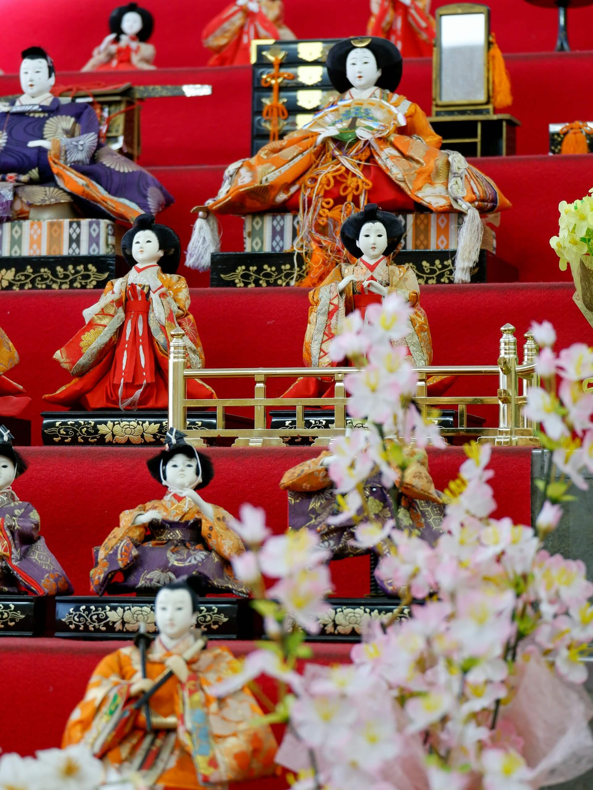 This image features a traditional display of Hina dolls used during the Doll Festival (Hina Matsuri) in Japan. The dolls are dressed in Heian period court attire and arranged on a multi-tiered red platform, symbolizing the emperor, empress, and their attendants.
