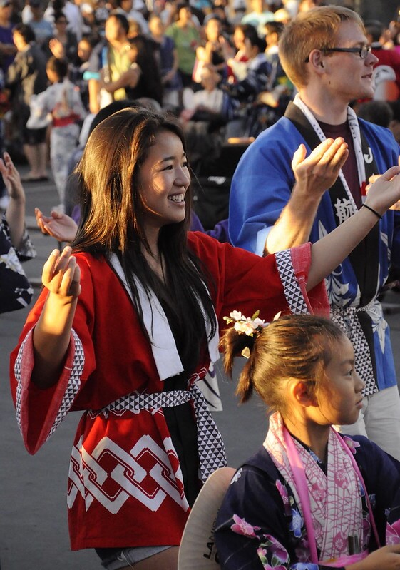 This image captures a joyful scene of a Bon Dance, a traditional Japanese dance event during the Obon Festival. The photo shows a young woman in a vibrant red and white yukata, smiling and dancing with her hands raised, surrounded by other participants in colorful attire, enjoying the communal festivities.
