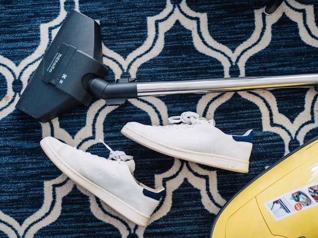 A vacuum cleaner and a pair of white sneakers placed on a patterned carpet, symbolizing the action of cleaning a room in preparation for someone's arrival, as expressed by the Japanese phrase "-teoku" in the sentence "彼女が来るから、部屋を掃除しておく" (I will clean the room because she is coming).
