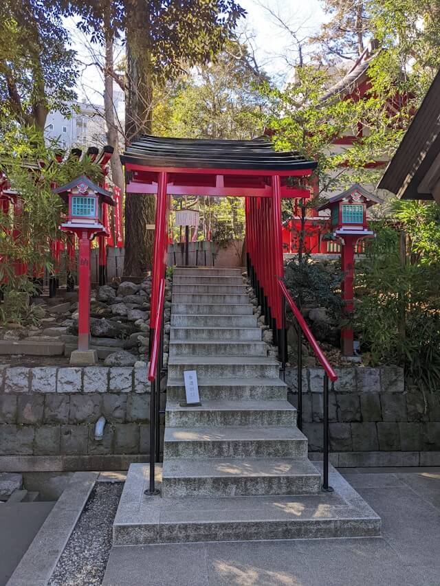 This image depicts a tranquil scene at a Shinto shrine, featuring a striking red torii gate at the top of a flight of stone steps, flanked by traditional lanterns and surrounded by lush greenery. The torii marks the entrance to the sacred space of the shrine, inviting visitors to enter and participate in the spiritual practices and rituals held within this serene setting.
